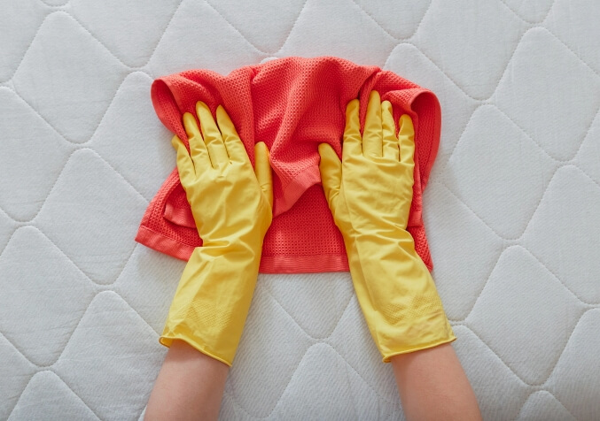 Two hands with yellow gloves cleaning mattress using red cotton cloth